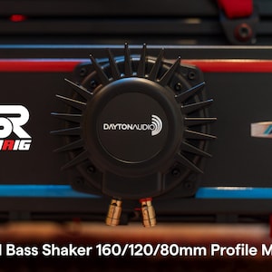 Puede incluir: Un shaker de bajos Dayton Audio BST-1 negro con montaje de perfil. El dispositivo tiene un diseño circular con un logotipo central y aletas radiales. La unidad está montada sobre una superficie negra con detalles en rojo y azul. El texto "SINHAIG" es visible.