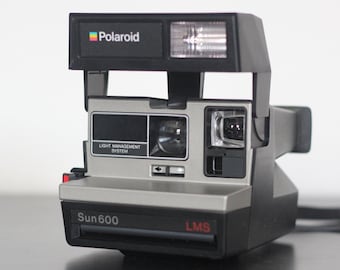 May include: A silver and black Polaroid Sun 600 instant camera with a black strap. The camera has a built-in flash and a light management system.