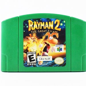May include: A green Nintendo 64 game cartridge for "Rayman 2: The Great Escape." The game's label features the title in yellow, a cartoon character, and the ESRB rating "E" for Everyone. The Ubi Soft logo is also visible.