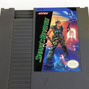 May include: A gray Nintendo Entertainment System (NES) game cartridge for Snake's Revenge, an Ultra Games release. The label features a green and black illustration of a soldier holding a gun and a knife. The text "Snake's Revenge" is in green and white, and the text "Ultra" is in white.