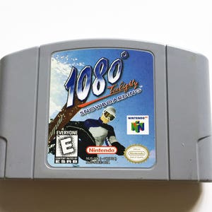 May include: A Nintendo 64 game cartridge for 1080° Snowboarding. The cartridge is gray with a blue and white label featuring a snowboarder in a blue and white suit. The label also includes the game title, "1080° Snowboarding", and the ESRB rating "Everyone".