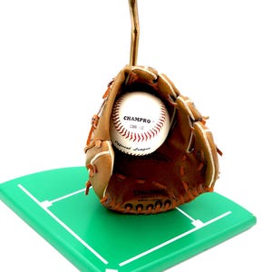 K&ouml;nnte beinhalten: Baseball-Themenlampe. Ein brauner Baseballhandschuh aus Leder h&auml;lt einen Baseball mit dem Text "CHAMPRO CBB 10 Official League". Die Lampe ist auf einer gr&uuml;nen Basis in Form eines Baseballfelds montiert.