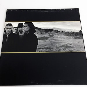 May include: Black and white album cover featuring a band of four men standing in front of a desert landscape. The album title "Three Joshuas" is printed in gold letters at the top of the cover.