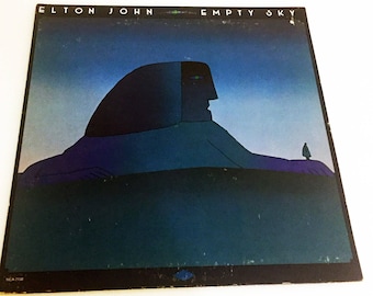 May include: A vintage vinyl record album cover featuring a blue and green illustration of a large, hooded figure with a single eye. The text "ELTON JOHN EMPTY SKY" is printed in white at the top of the cover.