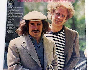 Puede incluir: Una portada de álbum de vinilo vintage con una foto de dos hombres, uno con una camisa a rayas y el otro con una chaqueta beige y un sombrero marrón. El título del álbum es "Simon and Garfunkel's Greatest Hits" y enumera las canciones incluidas en el disco.