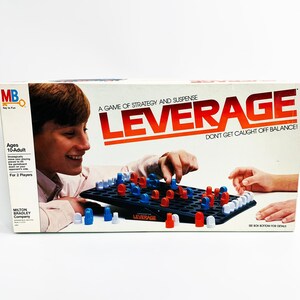 May include: A vintage board game box for "Leverage" by Milton Bradley. The box features the game's title in red and white, with the tagline "Don't Get Caught Off Balance!". The game board shows red and blue pieces. The box is designed for ages 10-Adult.