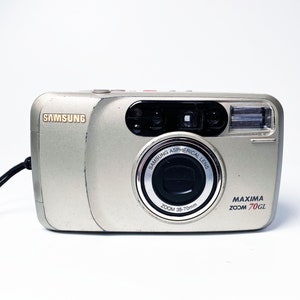 May include: A silver Samsung Maxima Zoom 70GL point-and-shoot camera with a black lens and a zoom range of 38-70mm.