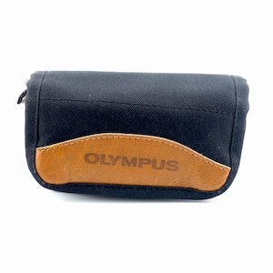 May include: A rectangular black fabric camera case featuring a tan leather-like accent panel on the front. The word OLYMPUS is embossed in the center of the tan panel. The case has a structured design with a zipper closure along the top edge.