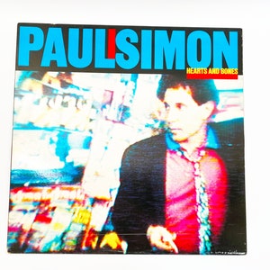 May include: A vintage vinyl record album cover featuring a blurry image of Paul Simon, the album title "Hearts and Bones" is printed in white text on a black background.