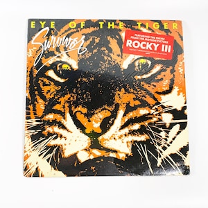May include: Album cover for Survivor's "Eye of the Tiger" featuring a tiger's face in orange and black. The album title and band name are displayed in white and orange. A red sticker indicates the theme song from the movie Rocky III.