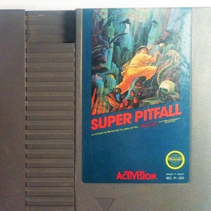 May include: A Nintendo Entertainment System (NES) game cartridge for Super Pitfall, a video game published by Activision. The cartridge is gray with a blue and white label featuring the game's title, a jungle scene, and the text "Licensed by Nintendo for play on the Nintendo Entertainment System."