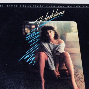 May include: A black vinyl record album cover with the title "Flashdance" in blue and white lettering. The cover features a woman in a grey sweater and black heels, sitting on a wooden floor with a red rug.