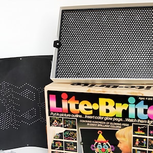 May include: A vintage Lite-Brite toy with a black plastic screen and a box with colorful images of designs that can be created with the toy. The box says "Lite-Brite" in rainbow letters and "Ages: 4 and Up".