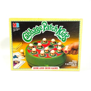 May include: A green and red game board with the words "Cabbage Patch Kids" and "Hide-and-Seek Game". The board has 12 holes with white mushroom-shaped figures in each hole. One figure is being removed from the board by a hand.
