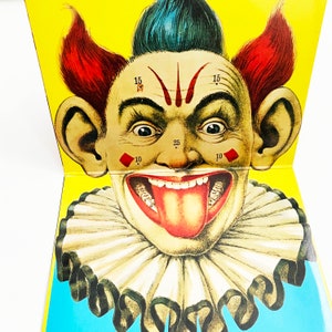 May include: A vintage carnival game target featuring a clown with red hair, a blue hat, and a wide, toothy grin. The clown's face is marked with numbers and diamond shapes, suggesting a target for throwing darts or other projectiles.