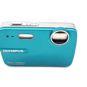 May include: A teal Olympus Stylus Water digital camera with a silver lens ring and a black wrist strap. The camera has a silver button on the top and the text "Olympus Stylus Water" is printed on the front.