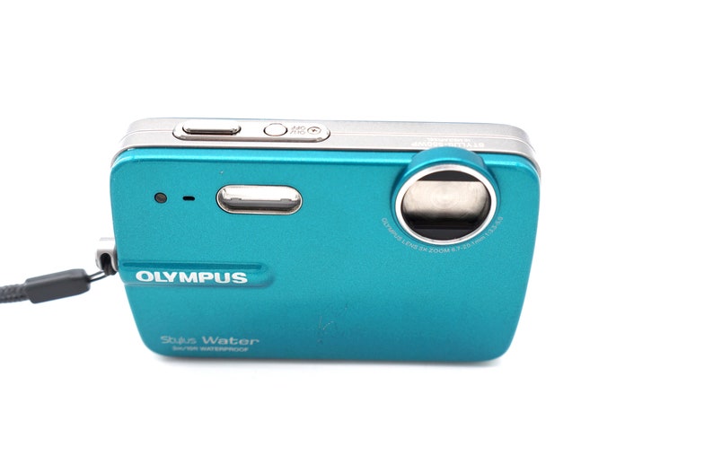 May include: A teal Olympus Stylus Water digital camera with a silver lens ring and a black wrist strap. The camera has a silver button on the top and the text "Olympus Stylus Water" is printed on the front.