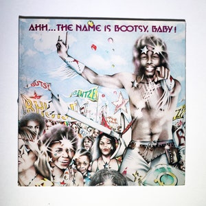 May include: A colorful album cover with a man wearing a white and red outfit and a star-shaped hat. The album cover has the text "AHH...THE NAME IS BOOTSY, BABY!" in large, bold letters.