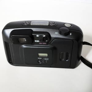 The back of a dark gray Pentax IQZoom 140 35mm film camera is shown, featuring the viewfinder, an LCD screen, and control buttons.