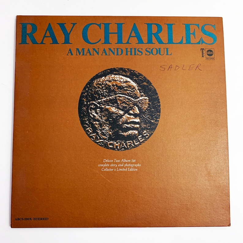 Pu&ograve; includere: Una copertina di album marrone con il testo "RAY CHARLES" in verde acqua e "A MAN AND HIS SOUL" sotto. Un'immagine circolare di Ray Charles &egrave; al centro. L'album &egrave; un Deluxe Two Album Set, edizione limitata.