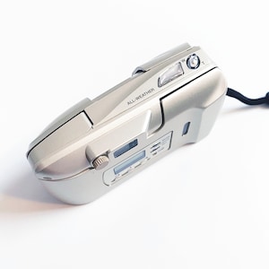 May include: A silver compact camera with a black strap. The camera has a digital display and the words "ALL-WEATHER" printed on the side.