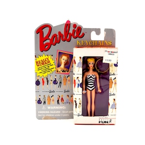 May include: A Barbie keychain in its original packaging. The doll is wearing a black and white striped swimsuit. The packaging includes the Barbie logo, the words "Keychains", and the text "Really Works!"