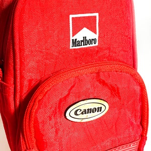 Op de afbeelding: Rode ritszak met een Marlboro-logo en een Canon-logo. De tas heeft een textuur aan de onderkant.