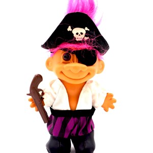 May include: A vintage troll doll dressed as a pirate, with a black hat featuring a skull and crossbones, an eye patch, and a toy pistol. The doll has pink hair and is wearing a white shirt and purple striped pants.