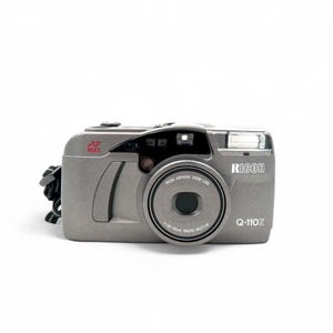 May include: A gray Ricoh Q-110Z camera with a zoom lens and flash. The camera has the text "RICOH" and "Q-110Z" printed on the front. The lens has the text "1-38-110mm MACRO MULTI-AF". A black strap is attached to the side.