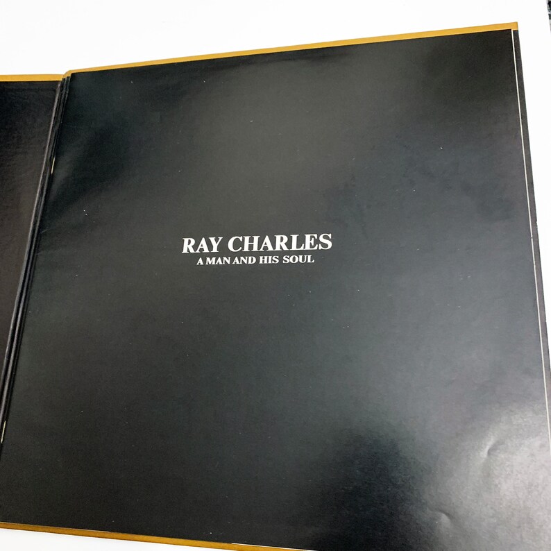 Pu&ograve; includere: Un libro aperto con copertina nera e bordi dorati. Il testo "RAY CHARLES A MAN AND HIS SOUL" &egrave; stampato in bianco. Le pagine interne del libro sono visibili.
