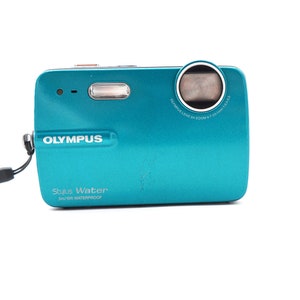 May include: A teal Olympus Stylus Water digital camera with a 3m/10ft waterproof rating. The camera has a black lens with a silver ring around it. The text "Olympus Stylus Water 3m/10ft Waterproof" is printed on the front of the camera.