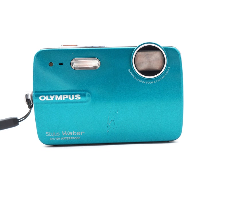 May include: A teal Olympus Stylus Water digital camera with a 3m/10ft waterproof rating. The camera has a black lens with a silver ring around it. The text "Olympus Stylus Water 3m/10ft Waterproof" is printed on the front of the camera.