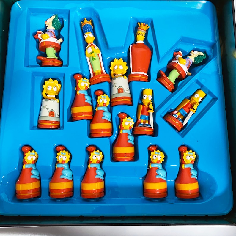 May include: A complete set of Simpsons chess pieces, featuring characters like Homer, Marge, Bart, Lisa, and Maggie. The pieces are made of plastic and are painted in bright colors. The chessboard is blue and has a grid pattern.