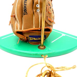 K&ouml;nnte beinhalten: Ein Baseballhandschuh mit dem Spalding-Logo liegt auf einer gr&uuml;nen, gebogenen Oberfl&auml;che mit wei&szlig;en Linien. Der Handschuh ist braun mit wei&szlig;en N&auml;hten, und eine goldfarbene Metallstange ist befestigt. Unten befindet sich eine goldfarbene Schnur.