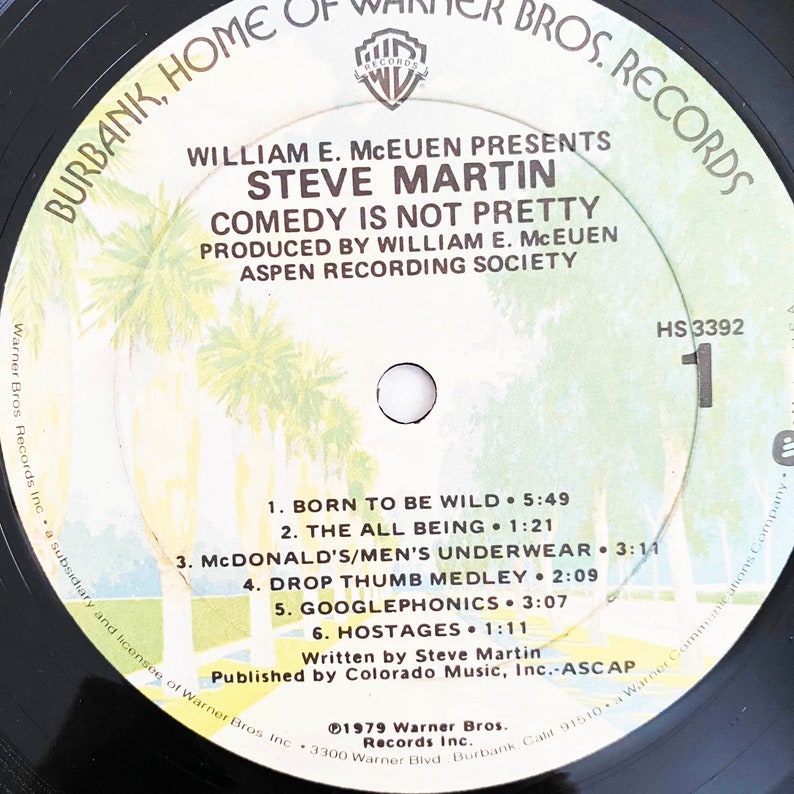 May include: A black vinyl record with a white label. The label features a palm tree design and text that reads "Burbank, Home of Warner Bros. Records", "William E. McEuen Presents Steve Martin", "Comedy Is Not Pretty", "Produced by William E. McEuen", "Aspen Recording Society", "HS 3392", "1", "Born to Be Wild", "The All Being", "McDonald's/Men's Underwear", "Drop Thumb Medley", "Googlephonics", "Hostages", "Written by Steve Martin", "Published by Colorado Music, Inc.-ASCAP", "1979 Warner Bros. Records Inc."