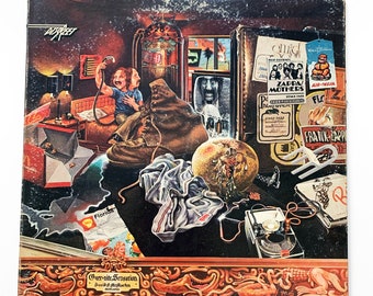 Puede incluir: Una portada de álbum de vinilo vintage que presenta una escena caótica con una bota, una fruta, una prenda de vestir y un hombre sosteniendo un micrófono. La portada del álbum es para "Over-Nite Sensation" de Frank Zappa y The Mothers of Invention.