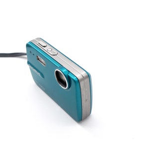 May include: A teal and silver digital camera with a lens cover and a strap.