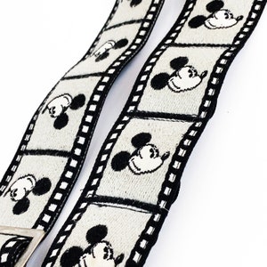 May include: A pair of black and white suspenders with a film strip design and Mickey Mouse heads. The suspenders are made of a woven fabric.
