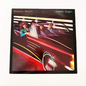 May include: A vintage vinyl record album cover featuring Bonnie Raitt with the title "Green Light" in white text. The cover art depicts a red car speeding down a road with yellow and blue lines, and a woman driving the car.