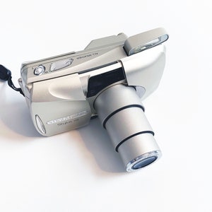 May include: A silver Olympus Stylus 120 point and shoot camera with a lens cap and a black strap. The camera is labeled "ALL-WEATHER" and has a zoom lens.