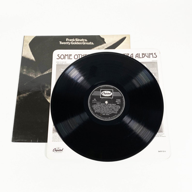 May include: A black vinyl record with a white label featuring Frank Sinatra's "Twenty Golden Greats" album. The record is on a white surface with a black and white album cover in the background. The album cover has the text "Frank Sinatra. Twenty Golden Greats. Some Other Capitol Albums."