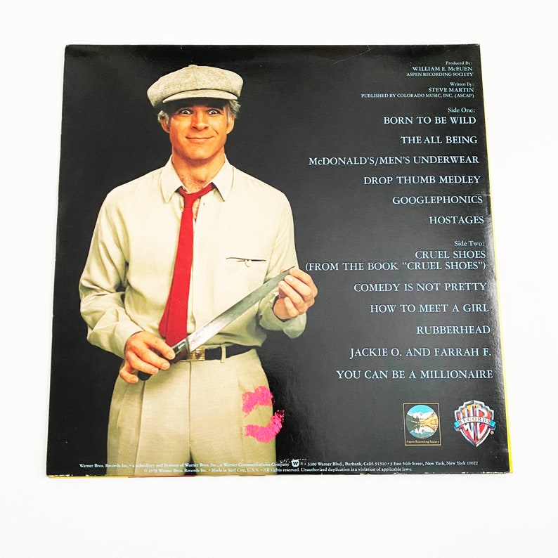 May include: A vinyl record album cover featuring Steve Martin in a white shirt, red tie, and a brown cap. He is holding a knife and has a serious expression on his face. The album cover is for Steve Martin's album "Cruel Shoes". The album cover is a black background with white text.