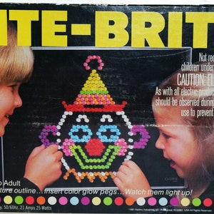May include: A Lite-Brite toy box with a clown image on the front. Two children are playing with the toy. The box says "Ages: 4 to Adult" and "Put in picture outline... insert color glow pegs... Watch them light up!"