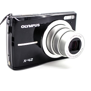May include: Black Olympus X-42 digital camera with a silver lens and a gray wrist strap. The camera has the Olympus logo and lens specifications printed on the lens. The camera is on a white background.