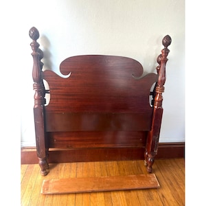 May include: A dark cherry-stained wooden headboard with a decorative, curved top and ornate, turned posts. The headboard has a rectangular base and a separate wooden slat. The headboard is a classic piece of bedroom furniture.
