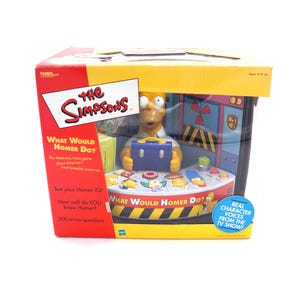 May include: A boxed "The Simpsons" electronic trivia game, featuring Homer Simpson holding a blue briefcase. The box is red and yellow with the game title "What Would Homer Do?" and includes text about trivia questions.