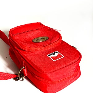 Op de afbeelding: Rode cameratas met een ritssluiting en een schouderband. De tas heeft een bruin leren patch met het woord "Canon" erop en een witte patch met het woord "Marlboro" erop.