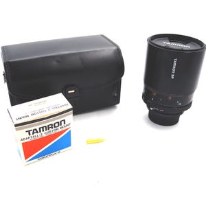 May include: A black Tamron SP camera lens with a black leather case and a box labeled "Tamron Adaptall-2 Custom Mount". The lens has measurement markings and the Tamron logo. A yellow cap is also present.