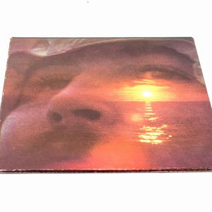 May include: A square music album cover featuring a double exposure photograph. The image overlays a close-up of a human face with a vibrant sunset over the ocean. The color palette includes deep purples, oranges, and yellows, with the sun reflecting on the water.
