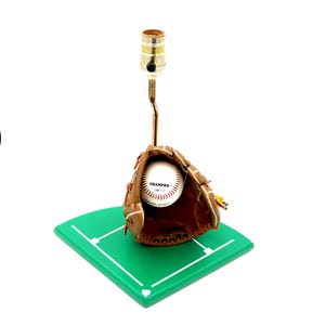 K&ouml;nnte beinhalten: Baseball-Themenlampe mit einem braunen Baseballhandschuh aus Leder, der einen Baseball h&auml;lt. Die Lampe hat einen messingfarbenen Hals und einen zylindrischen Lampenhalter. Der Sockel ist ein gr&uuml;nes, rautenf&ouml;rmiges Feld.
